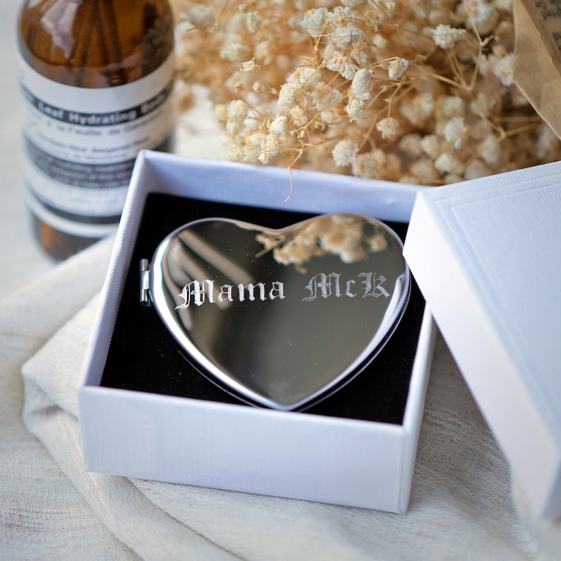 May include: A heart-shaped, silver compact mirror with the engraved text "Mama McK" inside a white gift box with a black interior. The mirror is reflective, and the box is open, revealing the mirror. A brown bottle and dried flowers are in the background.