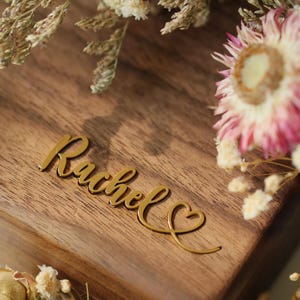 May include: A wooden box with the name "Rachel" in gold cursive script. The script includes a heart symbol. The box is surrounded by dried flowers with pink and white petals.