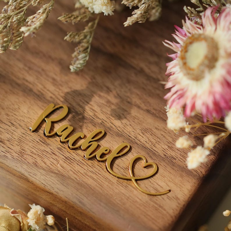 May include: A wooden box with the name "Rachel" in gold cursive script. The script includes a heart symbol. The box is surrounded by dried flowers with pink and white petals.
