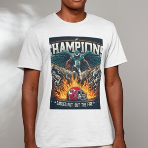 May include: A white t-shirt with a graphic of a football player in a green and white uniform with wings, surrounded by flames. The text "CHAMPIONS" is above the player and "EAGLES PUT OUT THE FIRE" is below the player.