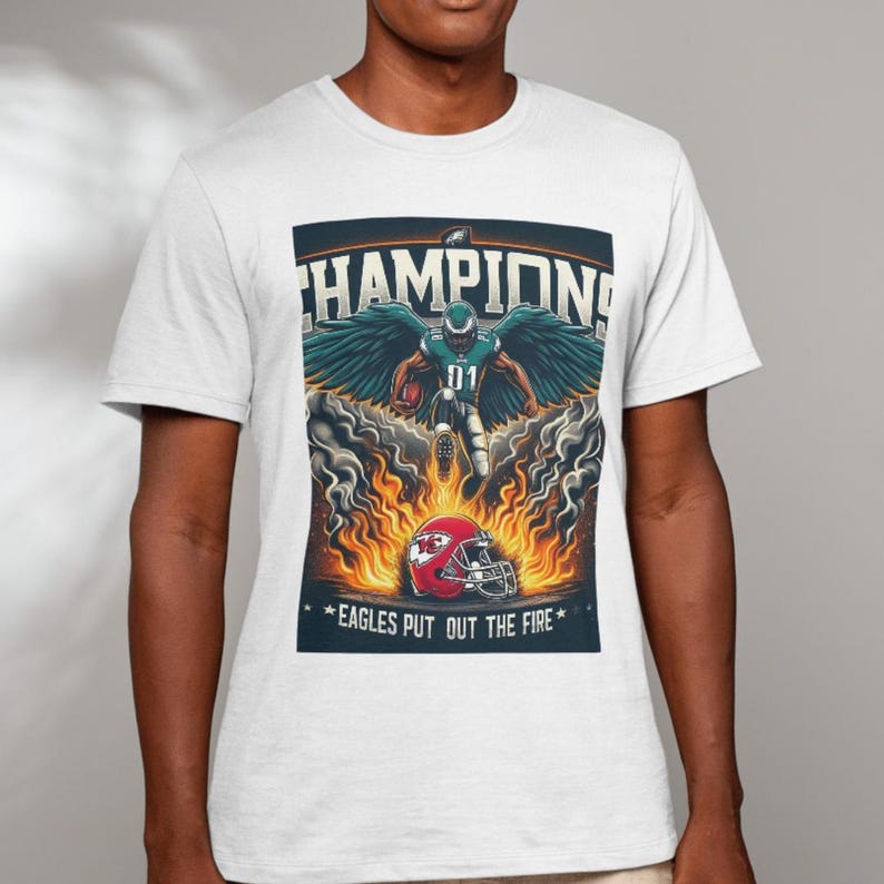 May include: A white t-shirt with a graphic of a football player in a green and white uniform with wings, surrounded by flames. The text "CHAMPIONS" is above the player and "EAGLES PUT OUT THE FIRE" is below the player.