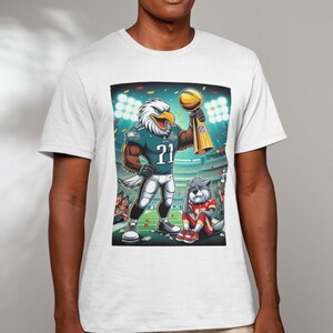May include: White t-shirt with a cartoon image of a Philadelphia Eagles mascot holding a trophy. The mascot is wearing a green and white jersey with the number 21. A cartoon dog mascot is sitting in the background.