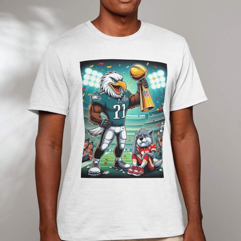 May include: White t-shirt with a cartoon image of a Philadelphia Eagles mascot holding a trophy. The mascot is wearing a green and white jersey with the number 21. A cartoon dog mascot is sitting in the background.
