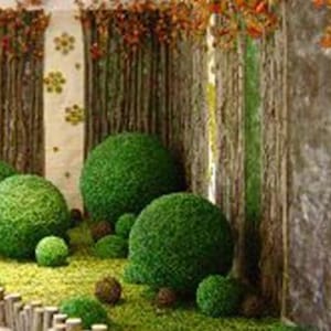 Może przedstawiać: Dekoracyjna ekspozycja z sztucznymi zielonymi kulami topiary o różnych rozmiarach, umieszczonymi pośród zielonej sztucznej trawy. Tło obejmuje sztuczne pnie i gałęzie drzew, tworząc leśną atmosferę. Brązowe drewniane ogrodzenie na pierwszym planie.
