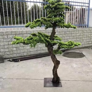 May include: A decorative artificial bonsai tree with a twisted, brown trunk and vibrant green foliage. The tree is mounted on a square, dark metal base. The background includes a brick wall and a metal fence.