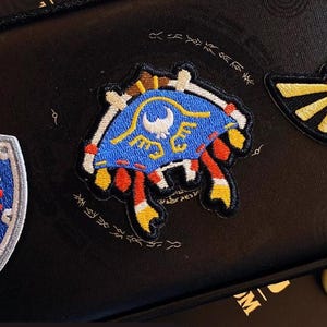 Puede incluir: Parches bordados con símbolos icónicos de la serie de videojuegos The Legend of Zelda. Incluye un escudo azul con un escudo rojo, una criatura azul y amarilla con forma de cangrejo y el símbolo Triforce dorado y negro.