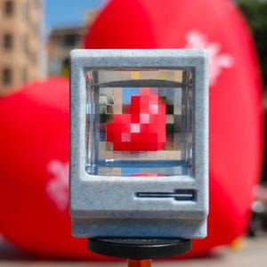 Puede incluir: Un pequeño objeto gris claro con forma de ordenador con un corazón rojo pixelado en su interior. El objeto está montado sobre un soporte negro y naranja, con un globo rojo en forma de corazón borroso en el fondo.