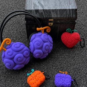 May include: A collection of silicone protective cases for headphones and earbuds, featuring purple, red, and orange fruit-inspired designs with spiral patterns and stems, accompanied by a dark wood-textured storage box with a metal latch.