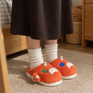 May include: A pair of orange plush slippers featuring a cartoonish creature design with white horns, a white snout, and circular green and blue accents on the sides. The slippers have a thick white sole and are worn with cream-colored ribbed socks.