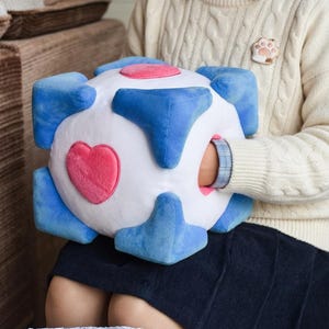 Puede incluir: Un juguete de cubo de peluche con base blanca, acentos azules en las esquinas y parches en forma de corazón rosa en los lados. El objeto suave y cuadrado tiene una abertura hueca en un lado, con una textura aterciopelada en toda la superficie.