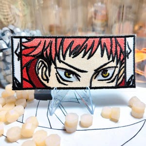 May include: Embroidered patch featuring a detailed anime-style illustration of a character's eyes and hair. The patch has a rectangular shape with a white border, red hair, and yellow eyes. The patch is displayed on a clear stand.