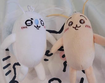 May include: Two plush toys, one white and one peach-colored, with cartoon faces and small arms and legs. The white toy has blue accents and closed eyes, while the peach toy has open eyes. Both have blush marks on their cheeks and small antennae.
