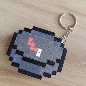May include: Pixelated grey keychain with a black border and a silver key ring. The design features red and white pixelated details. The keychain is a square shape and appears to be made of plastic.