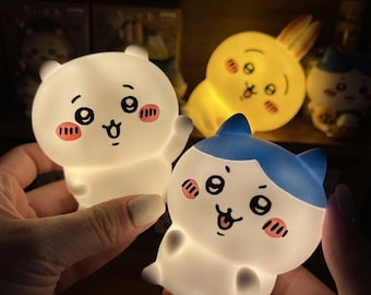 May include: Two small, glowing silicone night lights shaped like cartoon characters. One is a white bear-like figure with pink cheeks, and the other is a white cat-like figure with blue ears and pink cheeks. Both emit a soft, warm light.