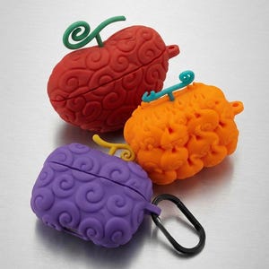 May include: Three silicone earbud cases shaped like stylised fruits with swirling, textured patterns. One case is red with a green stem, one is orange with a teal stem, and one is purple with a yellow stem and a black metal carabiner attached.