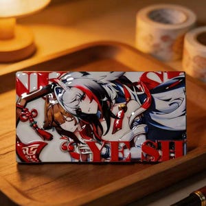 May include: A rectangular metal enamel pin featuring a detailed anime-style illustration of two characters with white and brown hair, red accents, and a small bell. The design includes bold red lettering that reads YE SHI across the bottom edge.