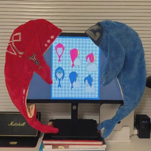 May include: A red and a blue plush toy shaped like a whale, flanking a computer monitor. The monitor displays a blue background with cartoon hair styles. A stack of books and a Marshall speaker are in the foreground.