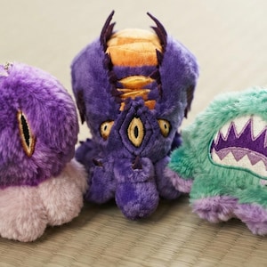 Puede incluir: Tres peluches de textura suave y afelpada. El juguete de la izquierda es morado con un solo ojo vertical. El central es morado con la parte superior naranja, múltiples ojos y apéndices tipo tentáculo. El de la derecha es de color verde azulado.