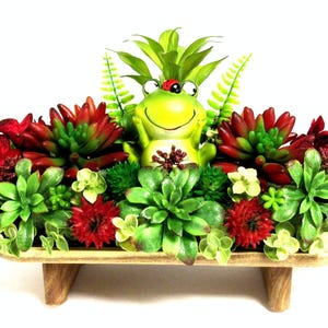 May include: A decorative arrangement featuring a central green frog figurine with a small ladybug on its head, surrounded by an assortment of artificial red and green succulents and ferns, all set within a rectangular wooden tray with four short legs.