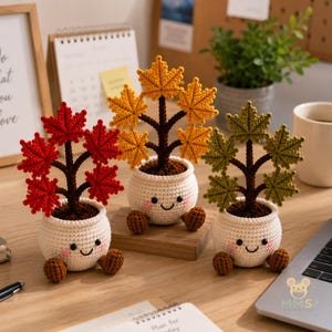 Pode incluir: Três plantas de crochê em vasos brancos sorridentes com pés castanhos e bochechas rosadas. Cada planta tem um caule castanho escuro e cinco folhas em forma de bordo em cores distintas: vermelho vibrante, amarelo dourado e verde-oliva.