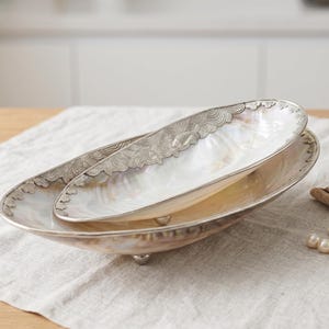 May include: Two oval serving dishes. The larger dish is silver-toned with three small feet. The smaller dish sits inside the larger one. Both dishes have a mother-of-pearl interior and a decorative silver-toned border.