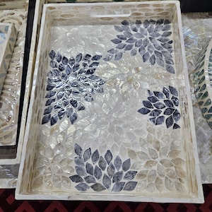 Pode incluir: Bandeja decorativa retangular com um design floral em mosaico. A bandeja apresenta um fundo branco com um padrão de folhas de cor clara e detalhes florais em azul escuro. A bandeja tem uma borda branca e um recorte de alça.