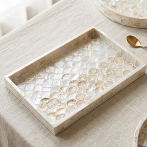 Könnte beinhalten: Rechteckige dekorative Schale mit einem Mosaik-Muscheldesign. Die Schale hat einen weißen Rand und eine Oberfläche aus schillernden, muschelförmigen Teilen in Weiß- und Beigetönen. Im Hintergrund ist ein goldener Löffel zu sehen.