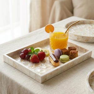 May include: A rectangular serving tray with a mother-of-pearl mosaic design holds a glass of orange juice with a gold straw, fruit, and pastries. The tray includes grapes, strawberries, kiwi, macarons, and small cakes. The tray is approximately 12 inches long.