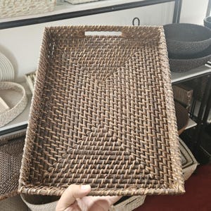 Puede incluir: Una bandeja rectangular de ratán tejido con un recorte para el asa. La bandeja es de color marrón natural y tiene un patrón tejido apretado. Otras cestas tejidas son visibles en el fondo.
