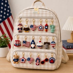 May include: A beige and white chequered backpack decorated with seventeen patriotic crochet keychains. The keychains feature designs including Uncle Sam, a bald eagle, hearts, stars, a rocket, the Statue of Liberty, a flag, a gnome, and a teddy bear.