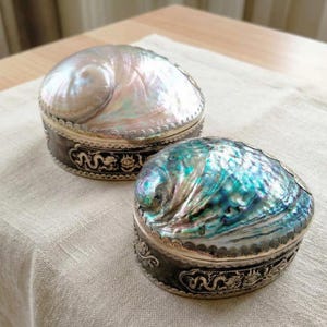 May include: Two decorative boxes with iridescent, pearlescent tops. The boxes have a silver-colored trim and dark gray sides with a decorative pattern. The boxes are round and sit on a light-colored surface.