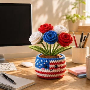 Puede incluir: Una maceta de ganchillo con una cara sonriente, decorada con la bandera estadounidense. Contiene flores de ganchillo rojas, blancas y azules. Las flores están dispuestas con hojas verdes, creando una exhibición floral patriótica.