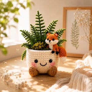 May include: A cream-coloured crocheted planter with a smiling face, pink cheeks, and two small brown feet. The planter holds a green crocheted fern and a small orange and white crocheted fox with black eyes, perched on the rim with its tail hanging down.