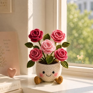 May include: A crocheted flower pot with a smiling face, holding several red and pink crocheted roses with green leaves. The pot is white with brown feet. The background includes a window and a book with the text "Every day holds little magic."