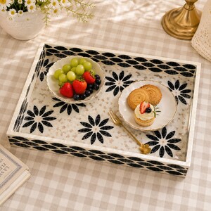 May include: Rectangular serving tray with a white background and black floral and geometric patterns. The tray holds small white plates with fruit and pastries. A gold fork rests on the tray. The tray is on a checkered tablecloth.