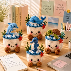 May include: Four crocheted containers with white bodies, smiling faces, and brown feet. Each features a different blue or green sea creature: a dolphin, a whale, a seahorse, and a turtle, all surrounded by crocheted seaweed, bubbles, and red starfish.