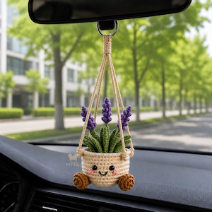May include: A crocheted hanging car ornament featuring a cream-colored pot with a smiling face, pink cheeks, and two small brown wheels at the base. The pot contains green crocheted leaves and three purple lavender stalks, suspended by cream cords.