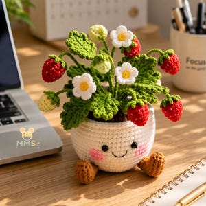 May include: A crocheted strawberry plant in a white pot with a smiling face, pink cheeks, and brown feet. The plant features green leaves, white flowers with yellow centers, and several red and pale green strawberries on thin stems.