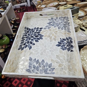 May include: A rectangular serving tray with a white frame and a mosaic floral design. The mosaic features a pattern of blue, beige, and white petal-shaped tiles. The tray has two handle cutouts.