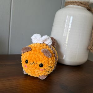 Op de afbeelding: Een handgemaakt, pluche, oranje en bruin gehaakt wezen met een lachend gezicht, zwarte ogen en een wit wolkachtig detail bovenop. Het speelgoed staat op een houten oppervlak.