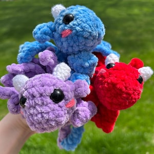 May include: Three handmade crochet axolotl plushies in blue, purple, and red. Each plushie has black safety eyes, white horns, and pink cheeks. The axolotls are soft and cuddly, perfect for a child's toy or a unique gift.