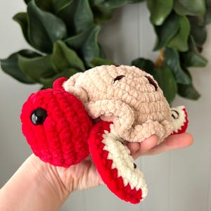May include: A handmade crochet turtle in shades of red and beige. The turtle has a red head with a black eye, a beige body, and red and white flippers. The turtle is a soft, plush toy.