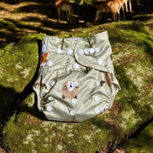 May include: A green and white reusable cloth diaper with a woodland animal print featuring a bear, deer, and fox. The diaper has white snaps and is folded in half.