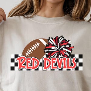 Puede incluir: Sudadera beige con un balón de fútbol y un diseño de pompón rojo y blanco. Las palabras "RED DEVILS" están impresas en rojo sobre un fondo a cuadros blanco y negro. La sudadera es de estilo casual.