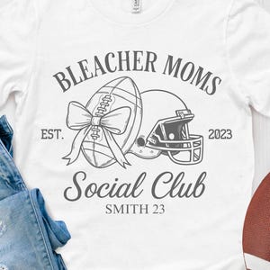 May include: White t-shirt with the text "Bleacher Moms Social Club" and a football graphic. The shirt is paired with blue jeans and a brown football. The design includes a football, helmet, and bow.