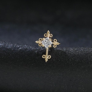 May include: A small, gold-coloured cross pendant with a central clear gemstone. The cross features ornate, swirling designs at the ends of each arm and along the vertical axis. The jewellery is set against a dark, textured background.