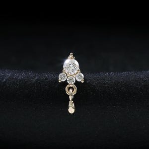 May include: A gold nose stud featuring a cluster of three small round clear stones arranged in a triangular pattern, with a single dangling gold teardrop-shaped charm suspended from a small circular ring at the base of the stone setting.