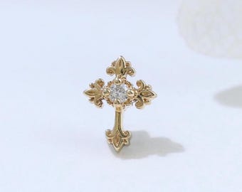 May include: A gold cross-shaped stud earring featuring an ornate, filigree-inspired design with decorative flourishes at each point. A single, small, clear round gemstone is set securely in the center of the cross, adding a subtle sparkle.