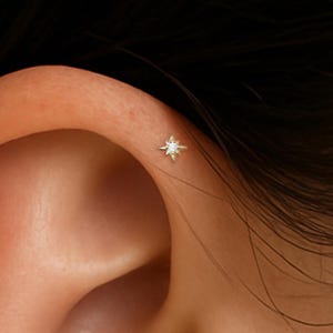 May include: Close-up of a person's ear with three small star-shaped earrings. The earrings are made of gold and have a single clear diamond in the center of each star.