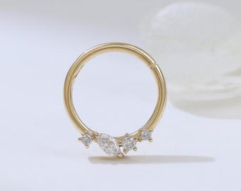 May include: A gold-colored hinged ring earring with a cluster of clear, sparkling gemstones. The gemstones are arranged in a delicate, curved design, adding a touch of elegance. The earring is set against a soft, white background.